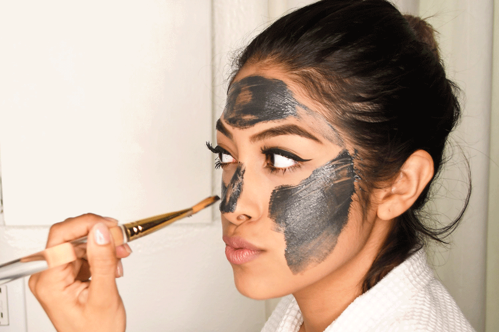 Woman applying black charcoal clay mask to face with brush