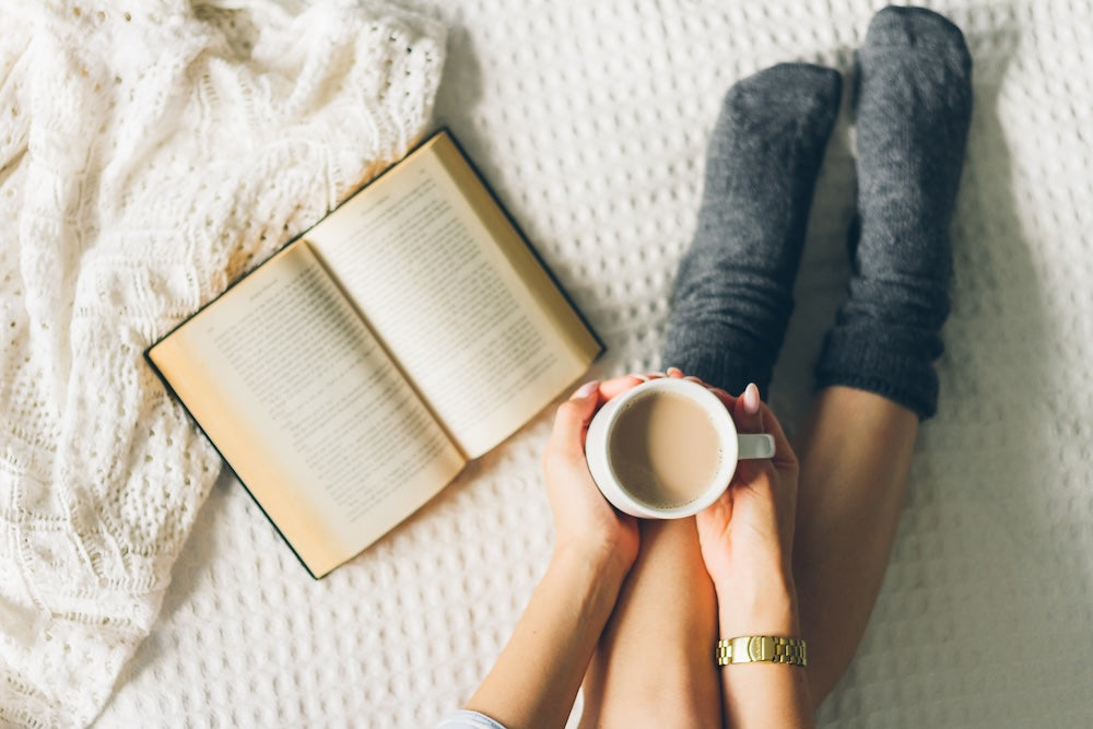 Woman in cozy socks holding coffee cup on a bed next to book and blanket