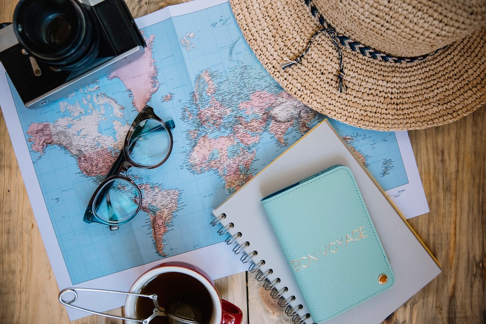 Travel Essentials scattered on table: glasses, notebook, hat, passport, and camera
