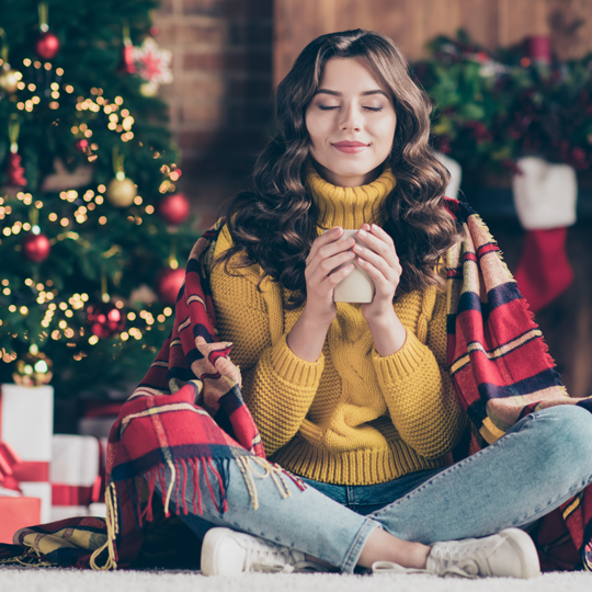 Woman in yellow turtleneck sweater holding warm drink near christmas tree and gifts