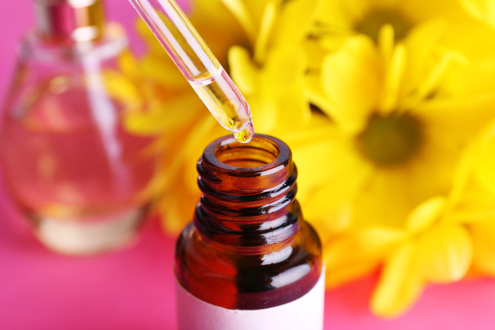 Chamomile Essential Oil in amber bottle with clear dropper, against pink background and yellow flowers
