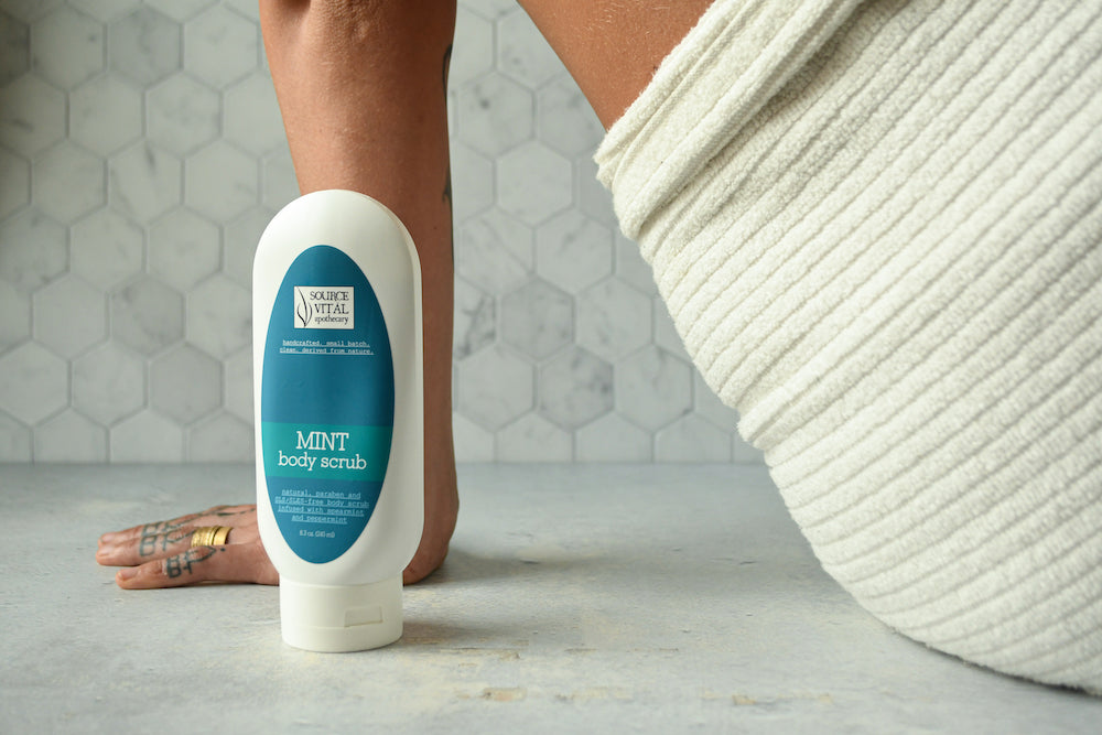 Woman in white towel leaning against marble floor with Mint Body Scrub bottle in foreground