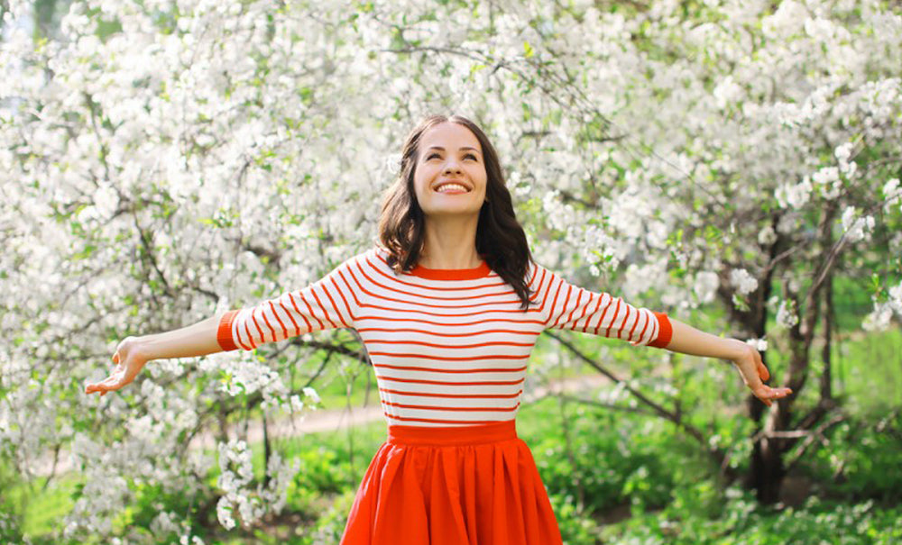Brunette woman with outstretched arms wearing white and red top, and red skirt against a background of cherry blossom trees
